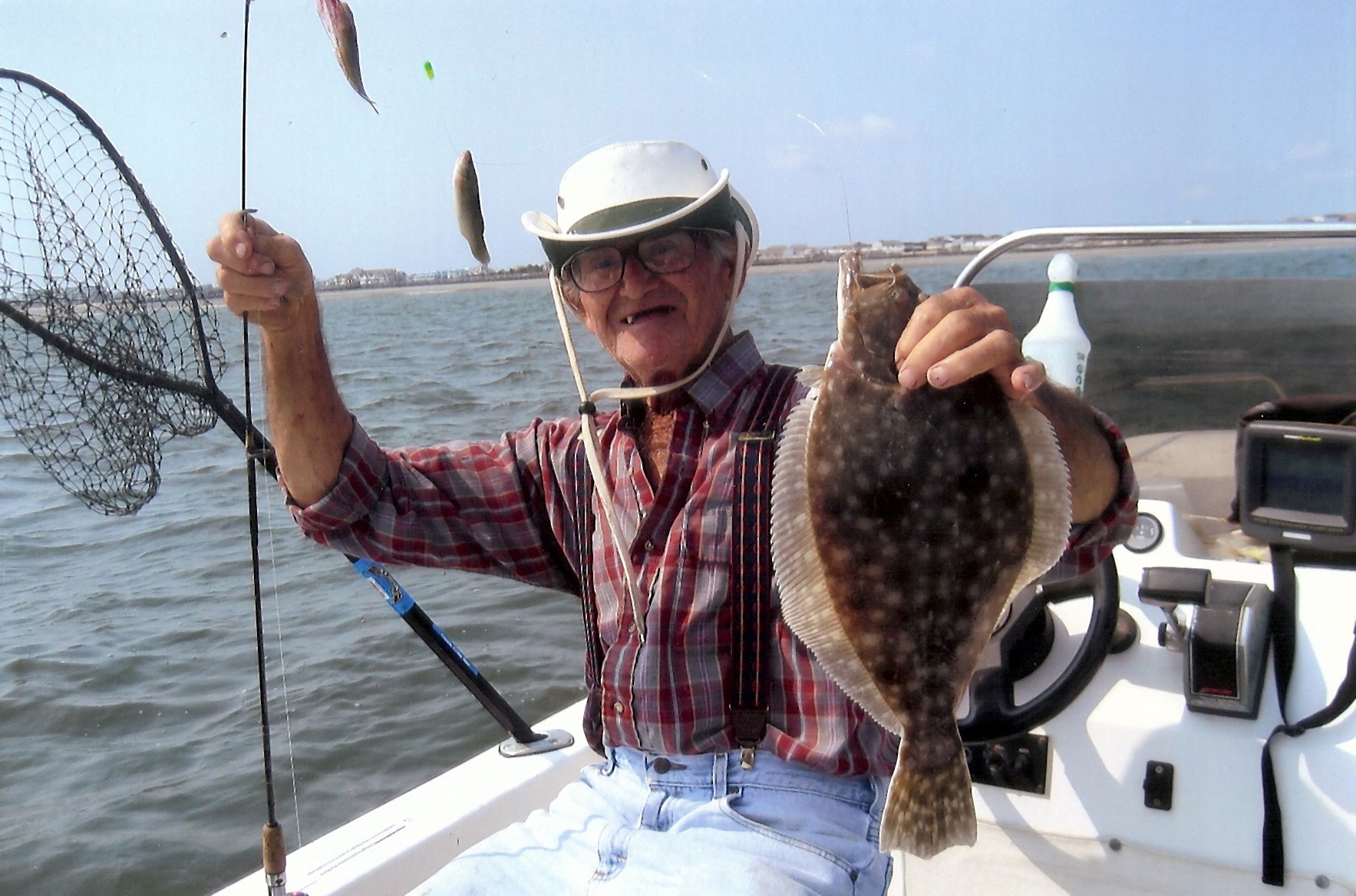 A smiling man in a white hat holds a flatfish and a fishing line on a boat